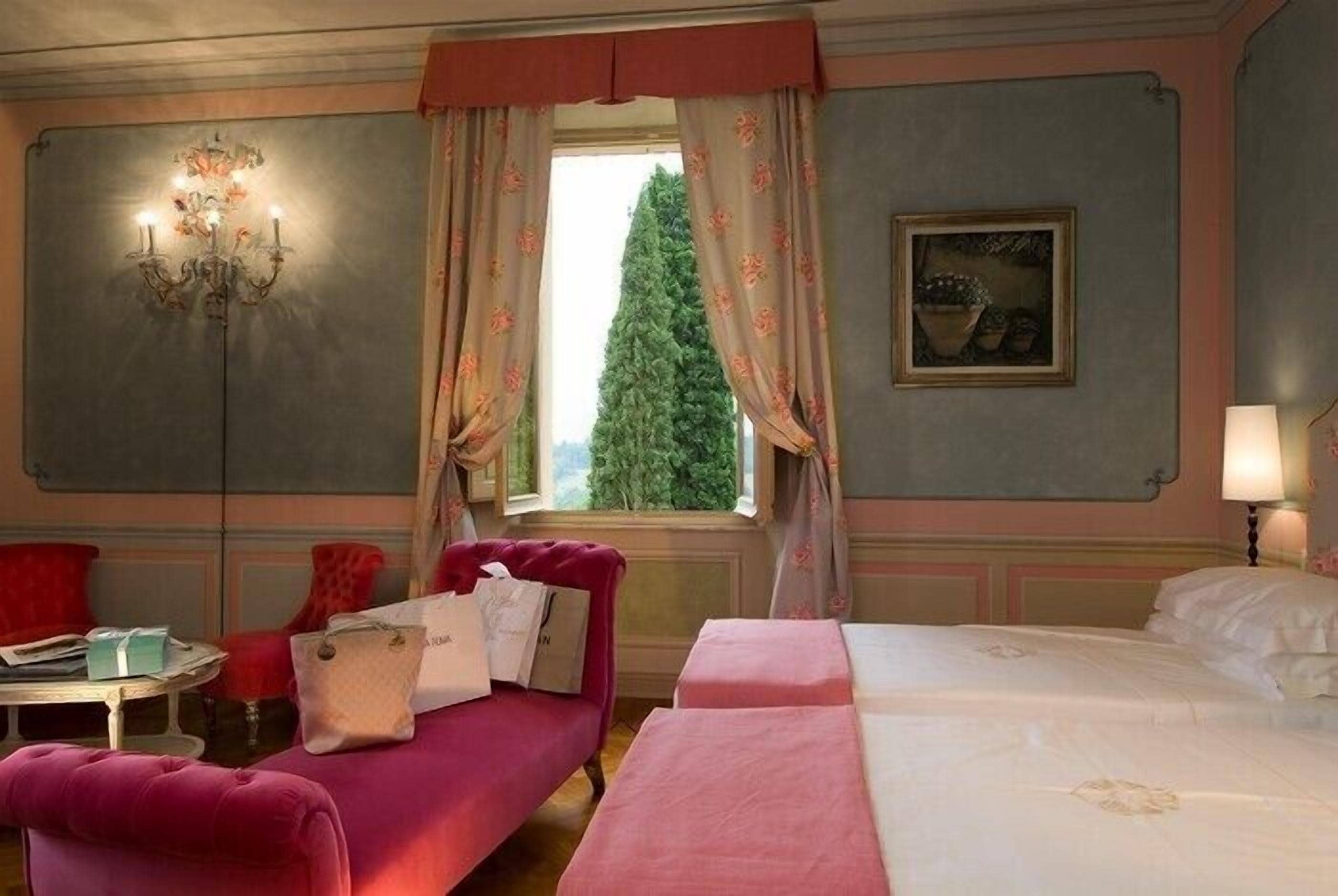Habitación de hotel con cama, sofá rojo, candelabro, ventana con cortinas y un cuadro en la pared.