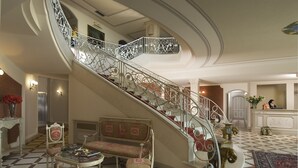 Un grand escalier orné de rampes en métal ouvragé, un sol à carreaux, un coin salon avec une table et, au fond, un comptoir d’accueil.