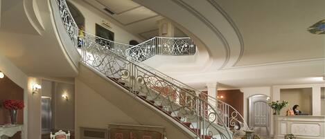 A grand staircase with ornate metal railings, a checkered floor, a seating area with a table, and a reception desk in the background.