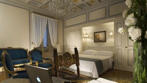 Une chambre d’hôtel avec un lit, un bureau avec un ordinateur portable, une chaise, un lustre et un vase rempli de fleurs.