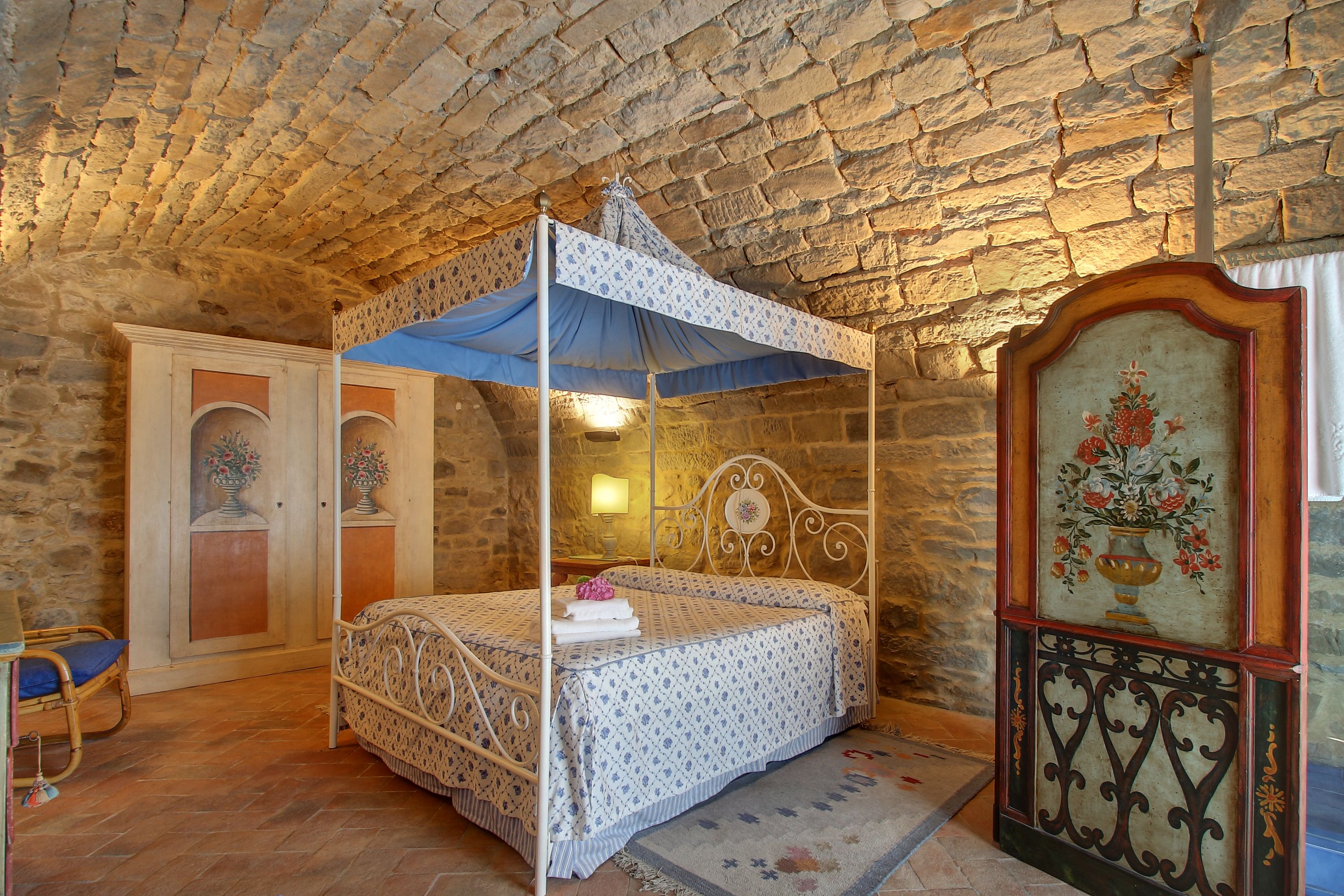 Un lit à baldaquin, une armoire en bois ornée d’une peinture florale et un mur en pierre.
