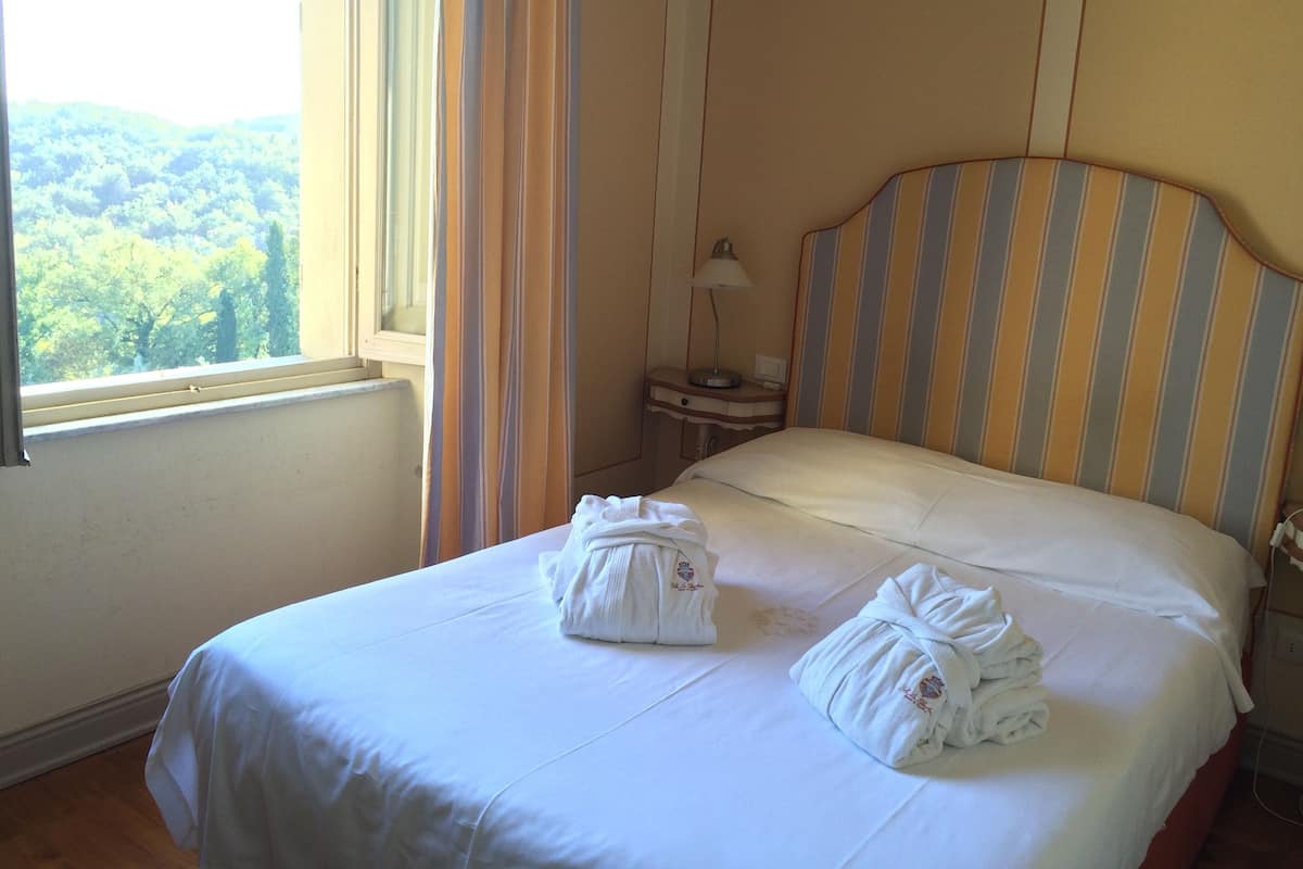 Una habitación de hotel con cama, mesita de noche, lámpara y ventana con cortinas.