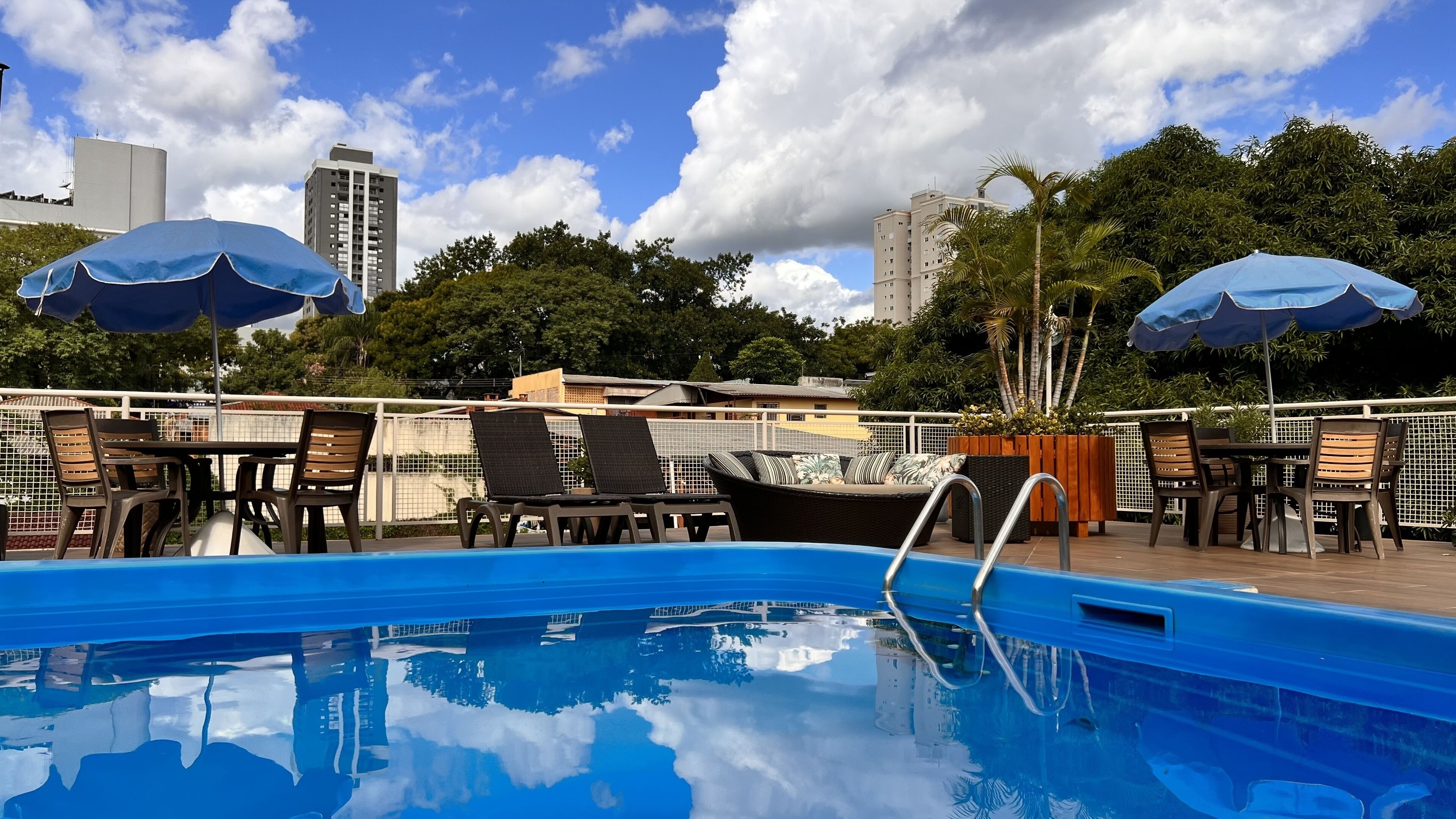 A rooftop pool area with lounge chairs and umbrellas, surrounded by a fence and trees, with a cityscape in the background.