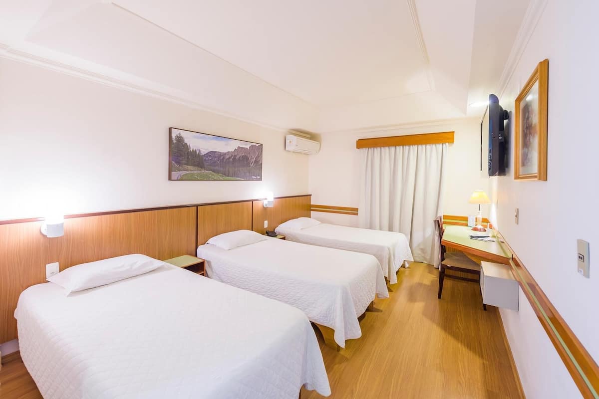 A hotel room with three single beds, a desk with a chair, a lamp, and a painting on the wall.