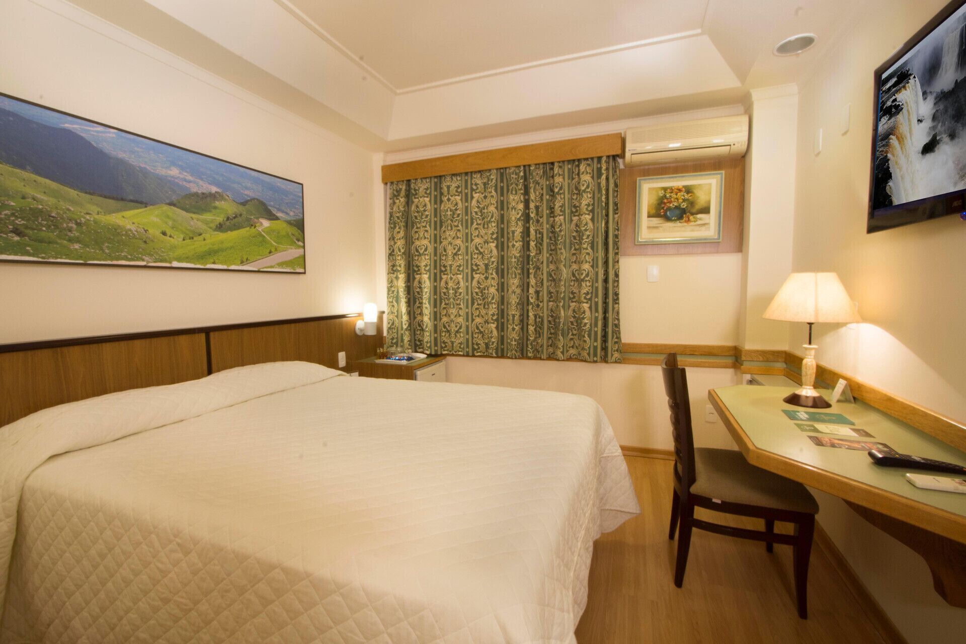 A hotel room with a bed, a desk, a chair, a window with curtains, a wall-mounted air conditioner, and a painting on the wall.