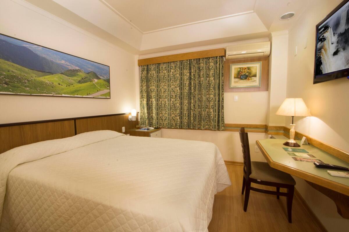 A hotel room with a bed, a desk, a chair, a window with curtains, a wall-mounted air conditioner, and a painting on the wall.
