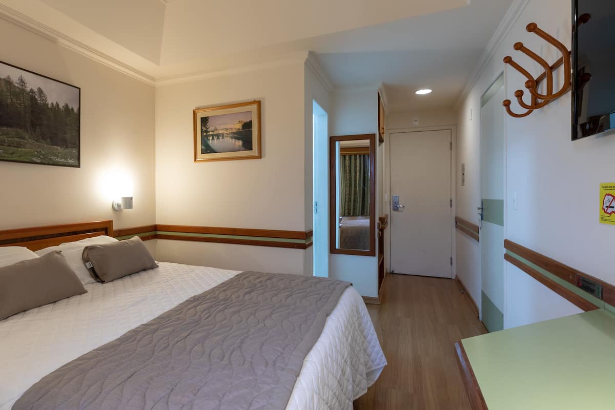 A hotel room with a bed, a painting, a door, and a coat rack.