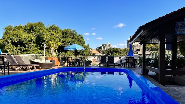 Una piscina con sillones de descanso y sombrillas, rodeada de vegetación y edificios.
