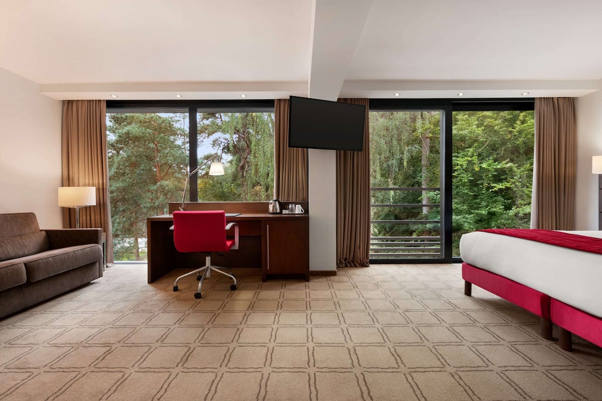 Une chambre d’hôtel équipée d’un lit, d’un bureau, d’une chaise, d’une télévision et de grandes fenêtres donnant sur la verdure.