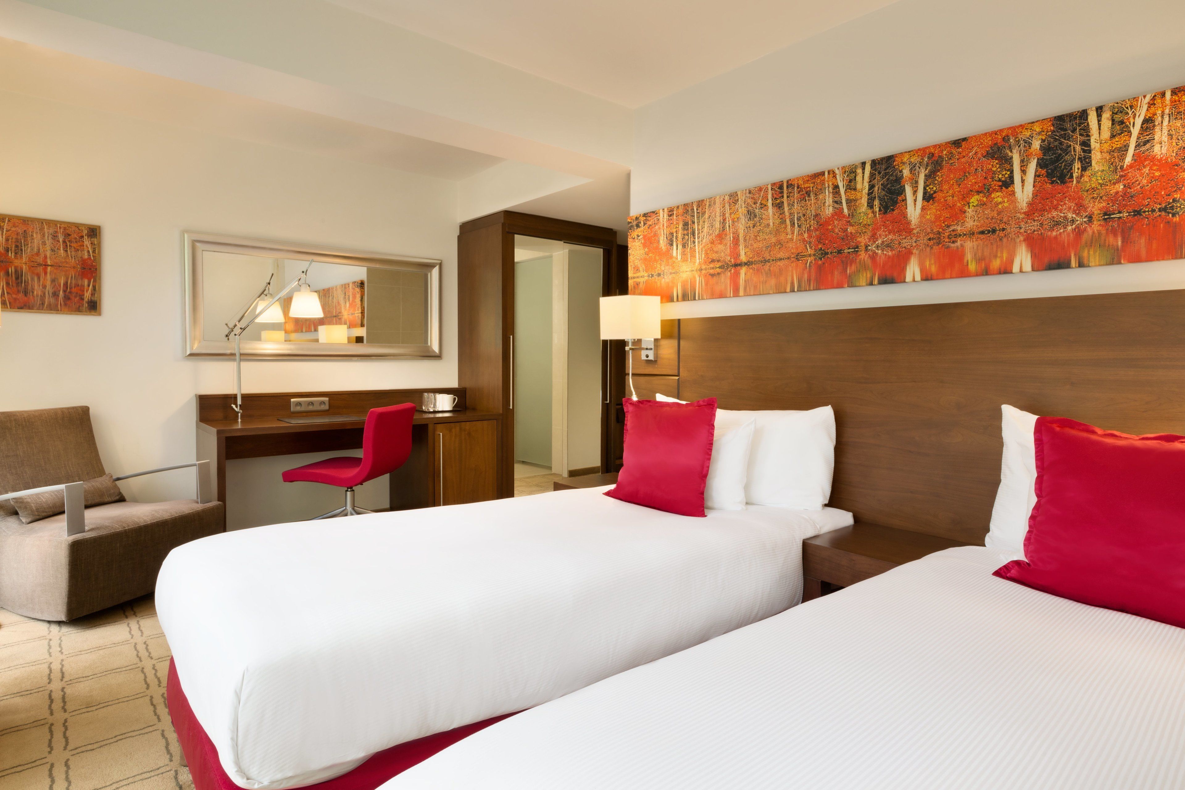 A hotel room with two beds, a desk, a chair, a mirror, and a painting on the wall.