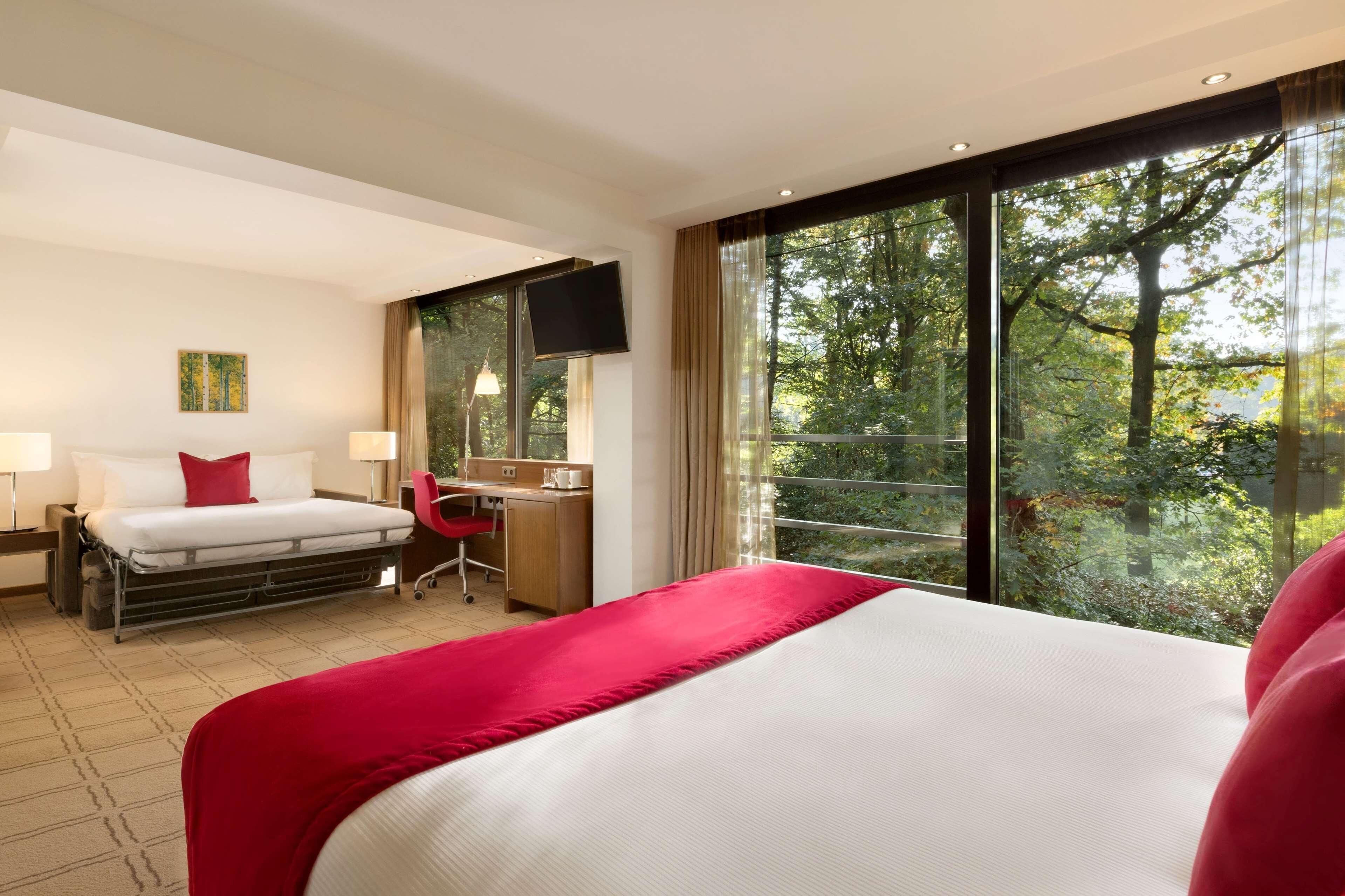 A hotel room with a bed, desk, chair, and large windows offering a view of trees.