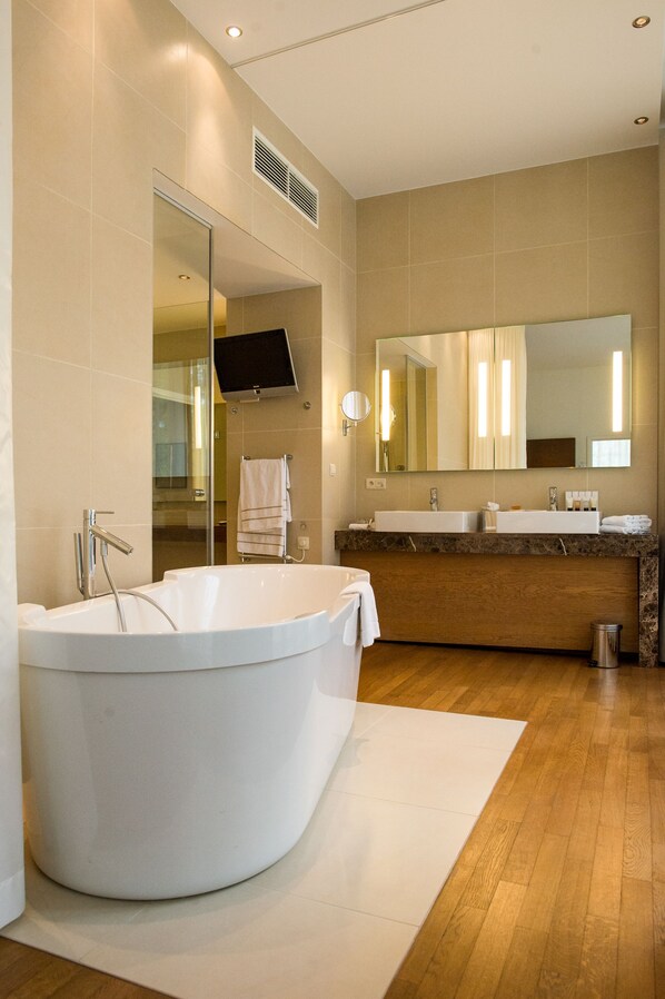 Un baño moderno con una bañera independiente grande, un tocador con doble lavabo, un espejo amplio y un televisor montado en la pared.