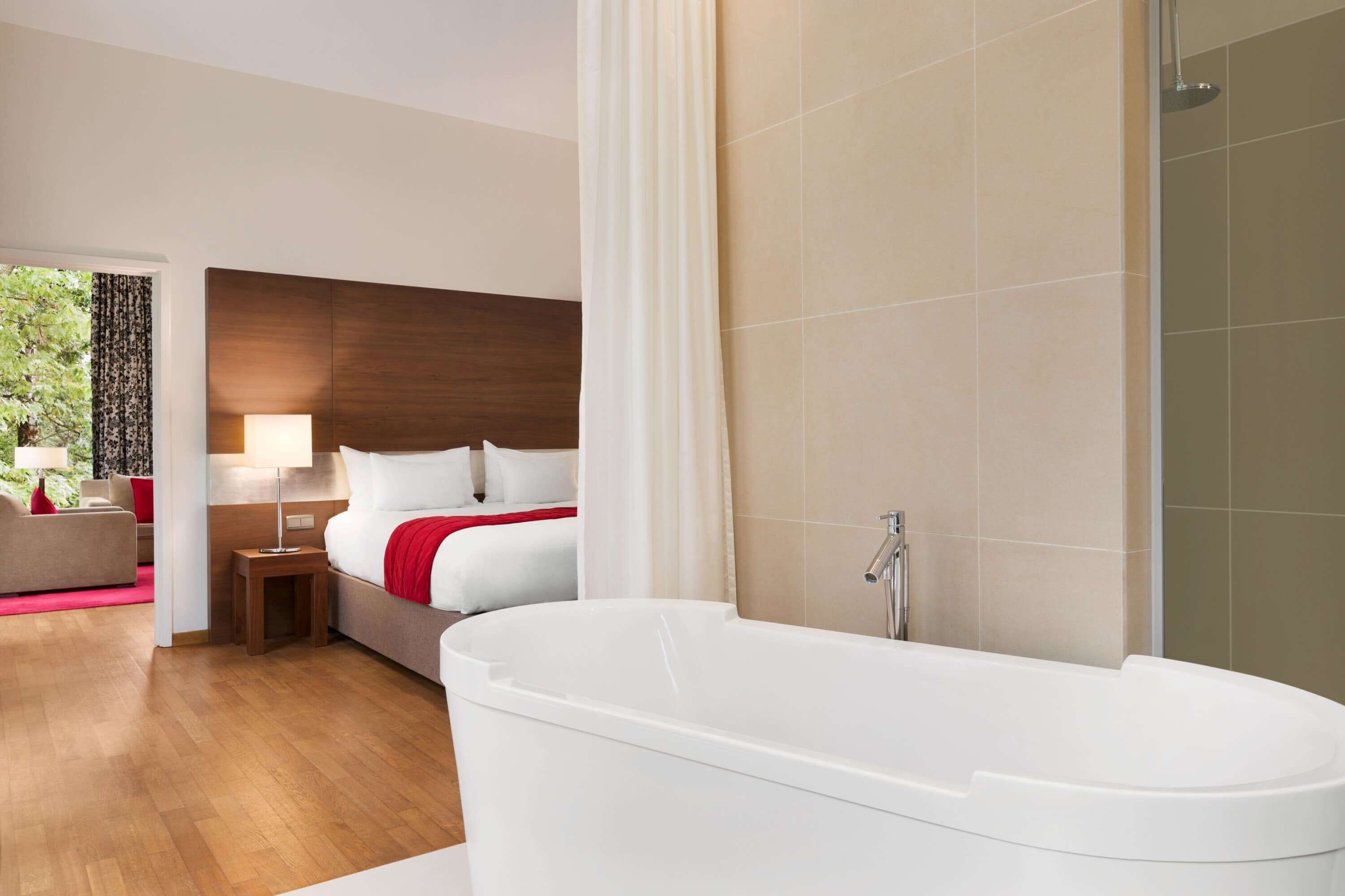 Habitación de hotel moderna con una bañera blanca grande, un cabecero de madera y vistas a zonas verdes.