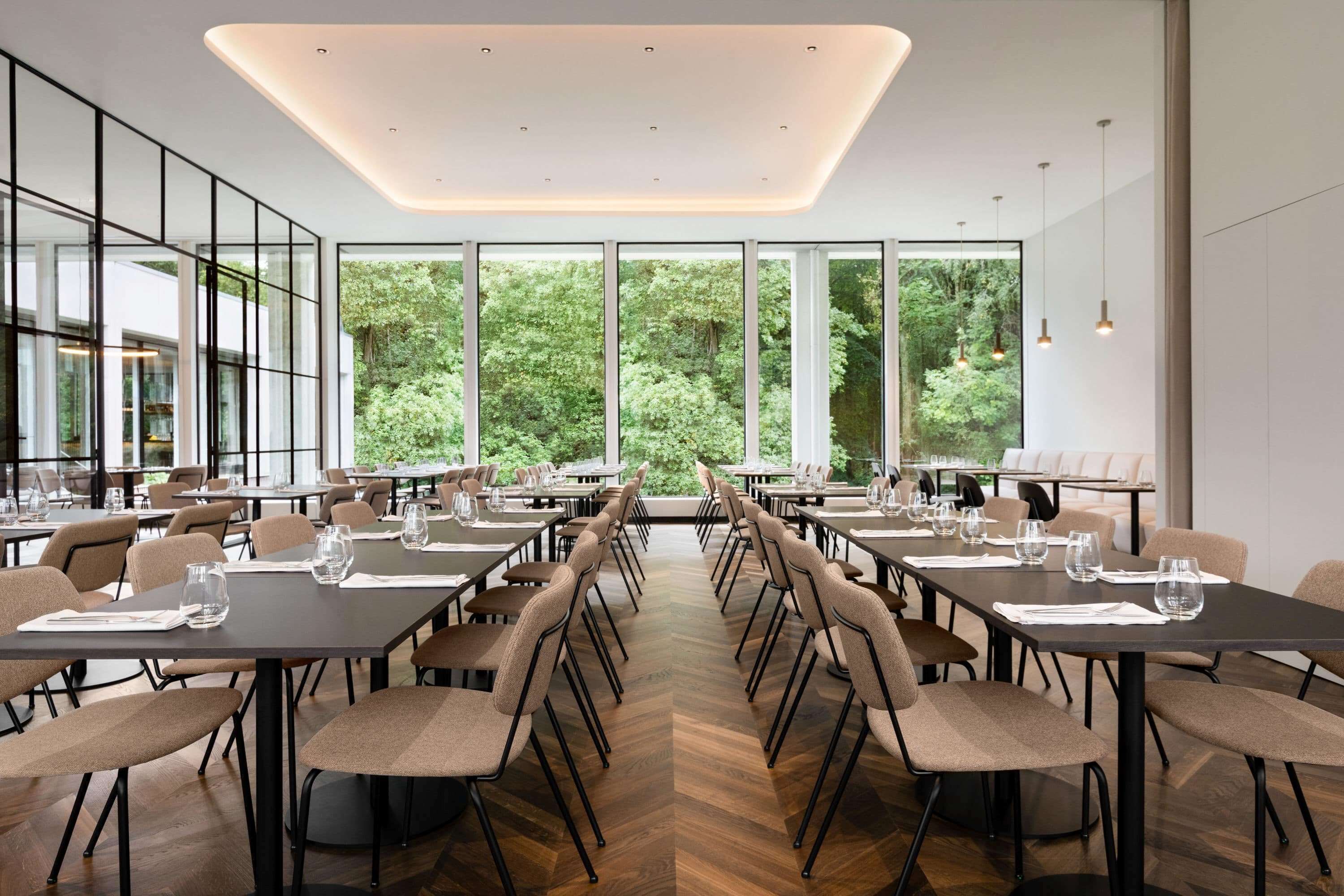Un espace repas moderne, doté de longues tables, de chaises et de grandes fenêtres offrant une vue sur la verdure.