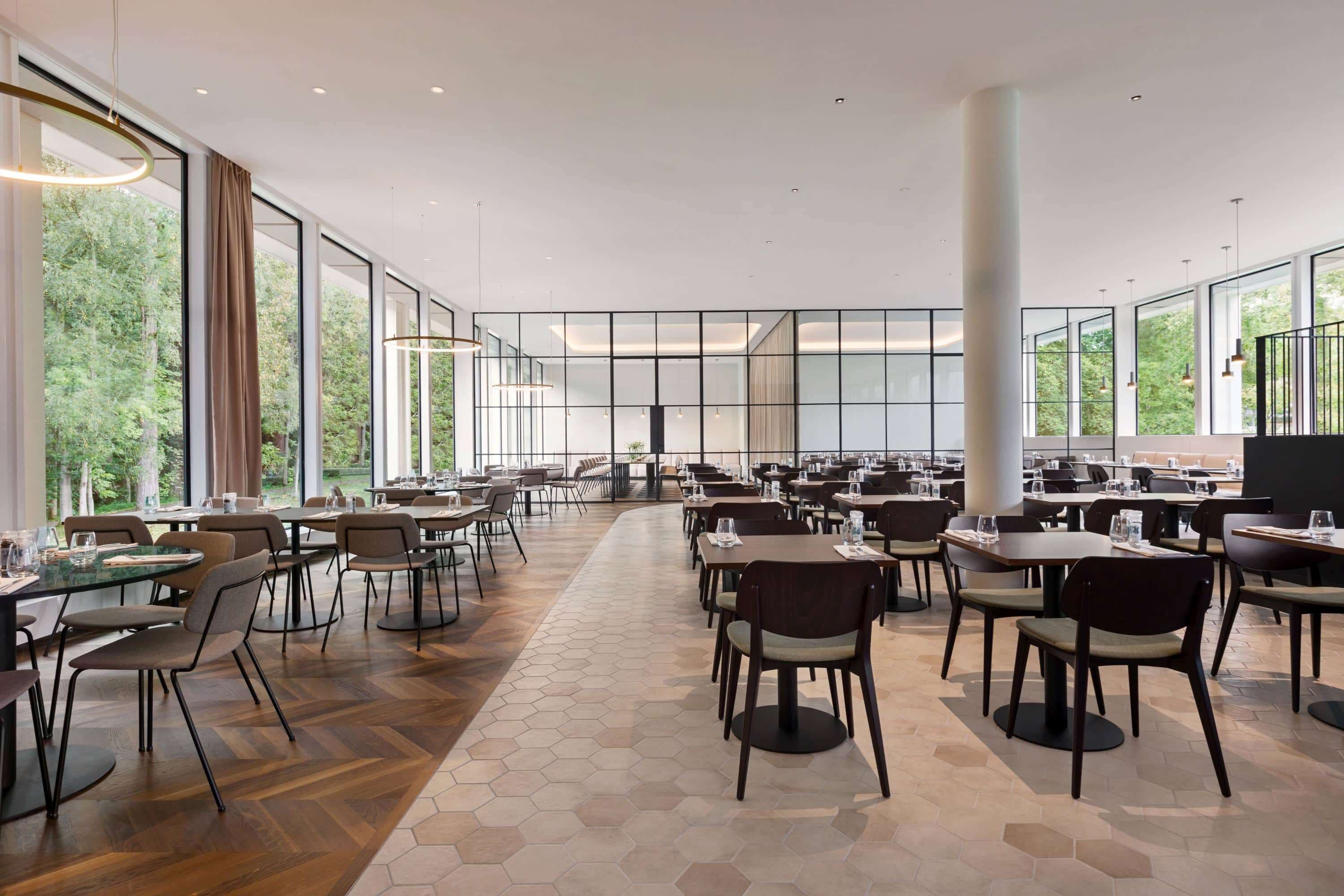 Un amplio comedor interior con múltiples mesas y sillas, grandes ventanales que ofrecen vistas a zonas verdes, y un diseño arquitectónico moderno con suelo de baldosas hexagonales.