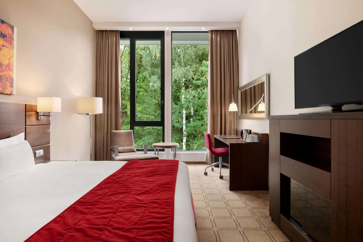 Une chambre d’hôtel avec un grand lit, un bureau, une chaise, une télévision et une vue sur les arbres par la fenêtre.