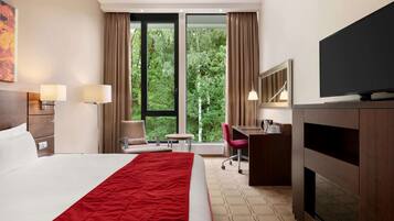 Een hotelkamer met een groot bed, een bureau, een stoel, een televisie en uitzicht op bomen.