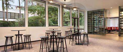 Un interior moderno con ventanas amplias, varias mesas y taburetes, y una estantería para vinos al fondo.