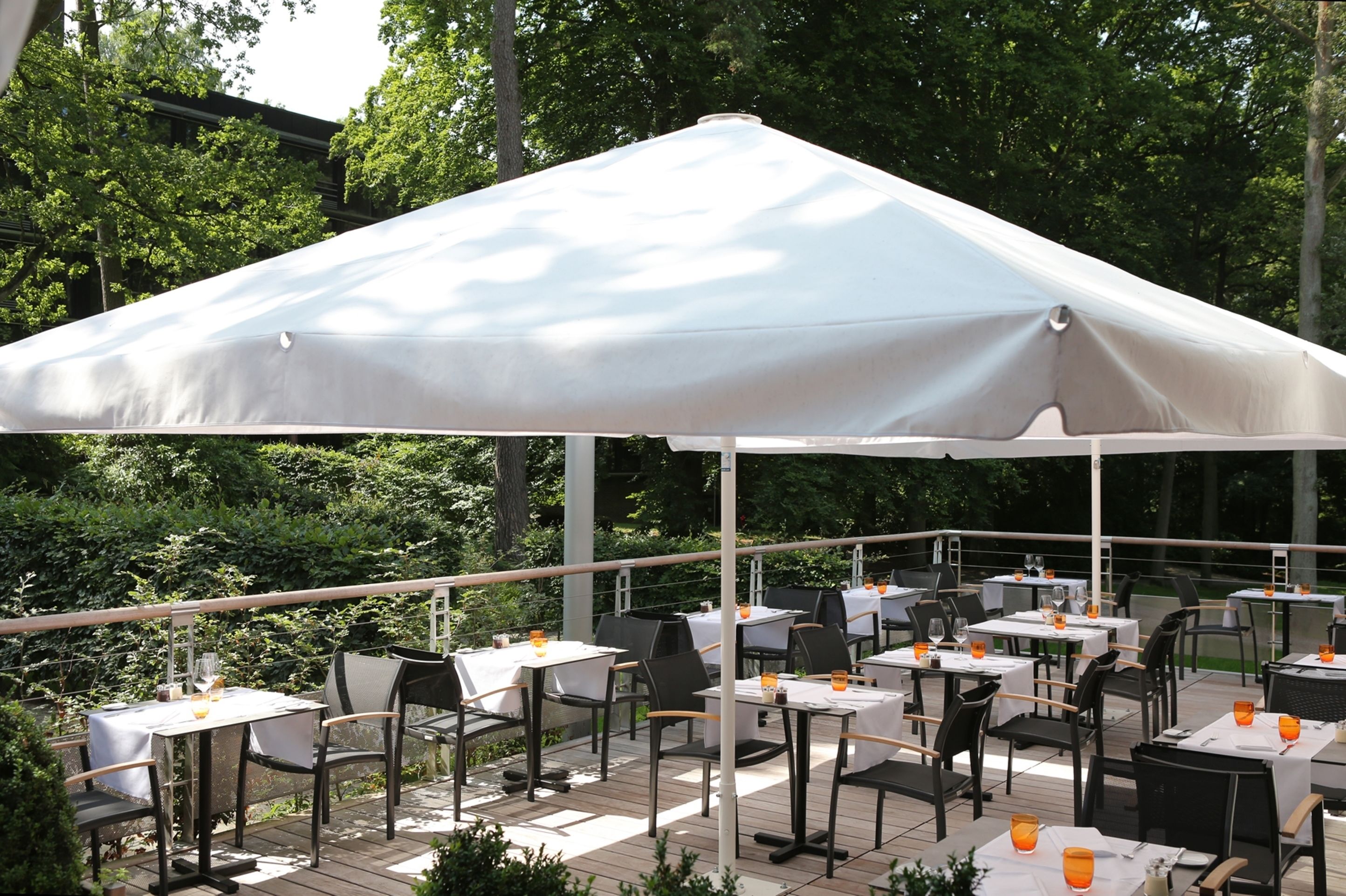 An outdoor patio with tables and chairs under a large umbrella, surrounded by greenery.