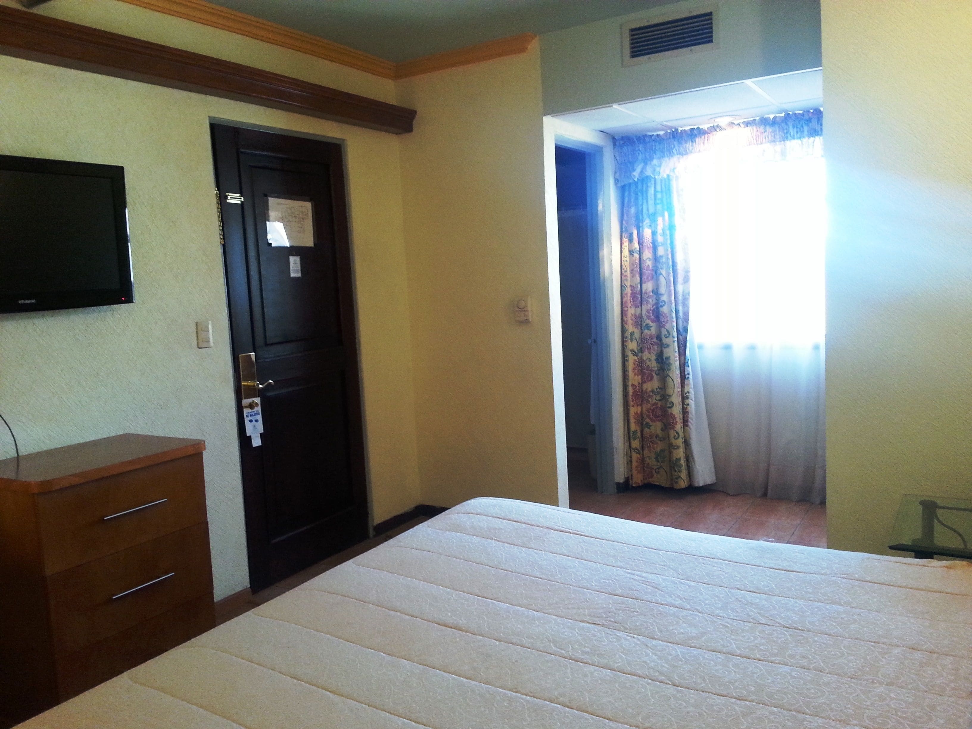 A hotel room with a bed, a wooden dresser, a television mounted on the wall, and a door with a lock.