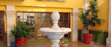 A courtyard with a central fountain, potted plants, and a yellow building with a window and balcony.