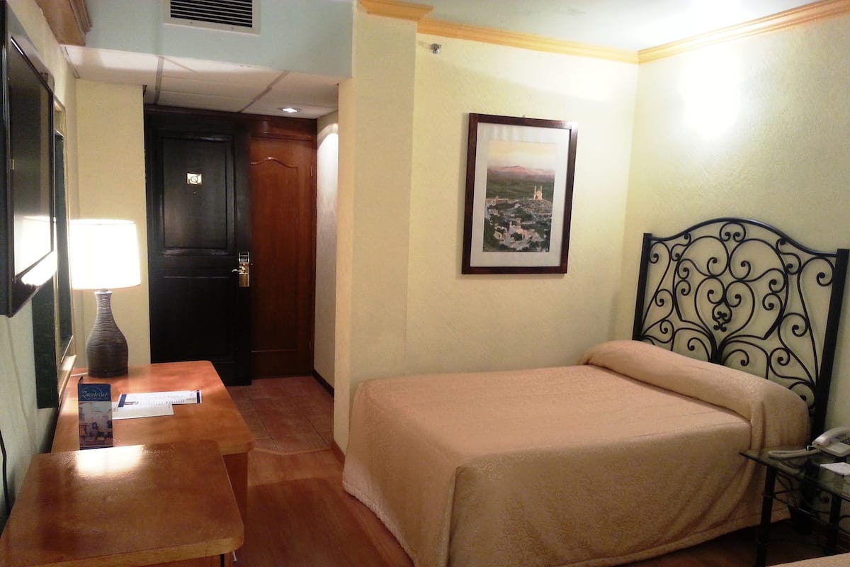 Een hotelkamer met een bed, een bureau met lamp, een ingelijst beeld aan de muur en een deur naar een andere kamer.