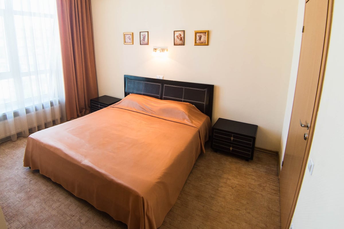 A bedroom with a large bed, two bedside tables, a nightstand, a window with curtains, and four framed pictures on the wall.