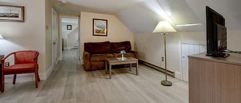 A room with a wooden floor, a brown sofa, a red chair, a small table, a floor lamp, and a television on a wooden cabinet.