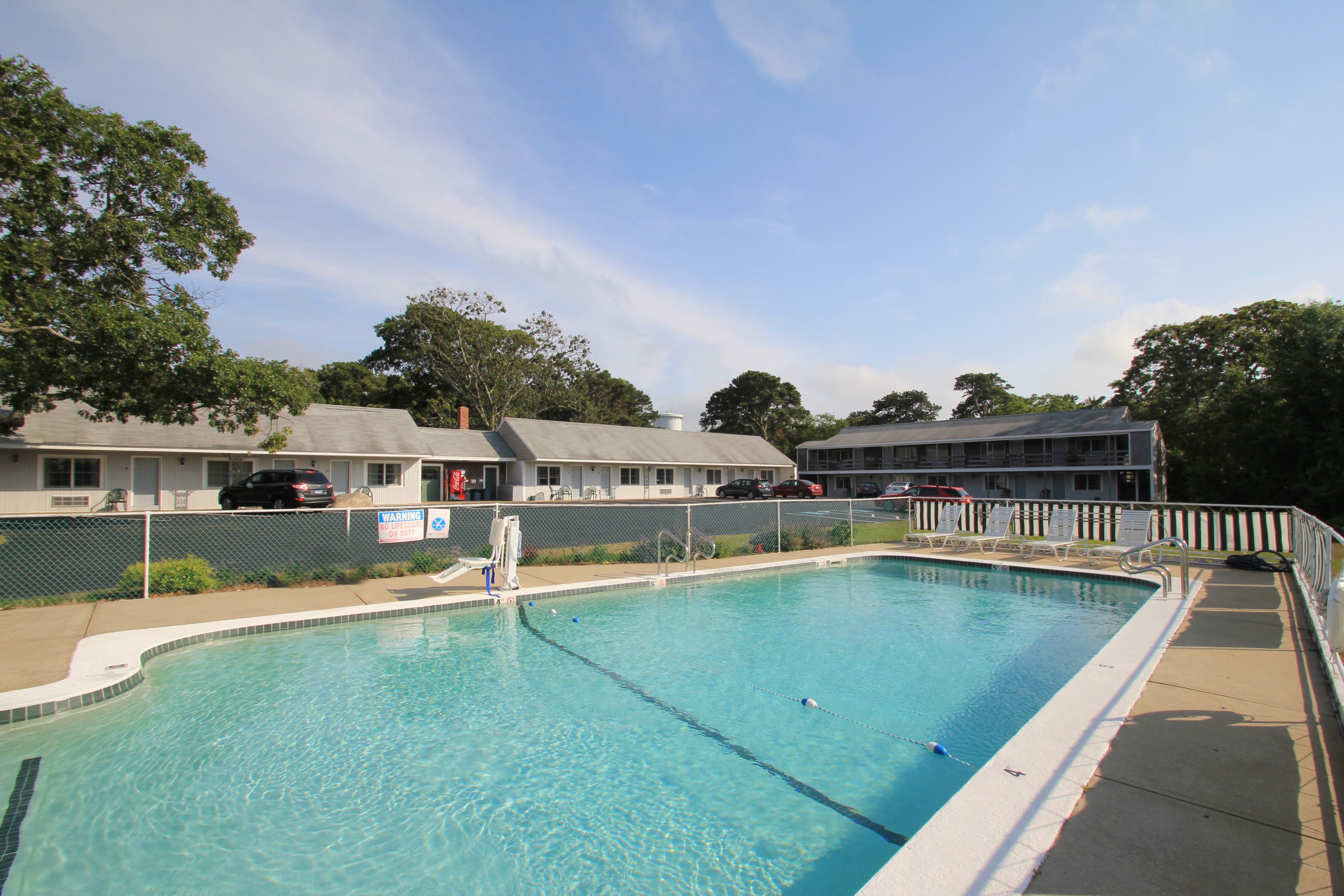 A swimming pool with a fenced deck, surrounded by a building with multiple rooms and a covered parking area.