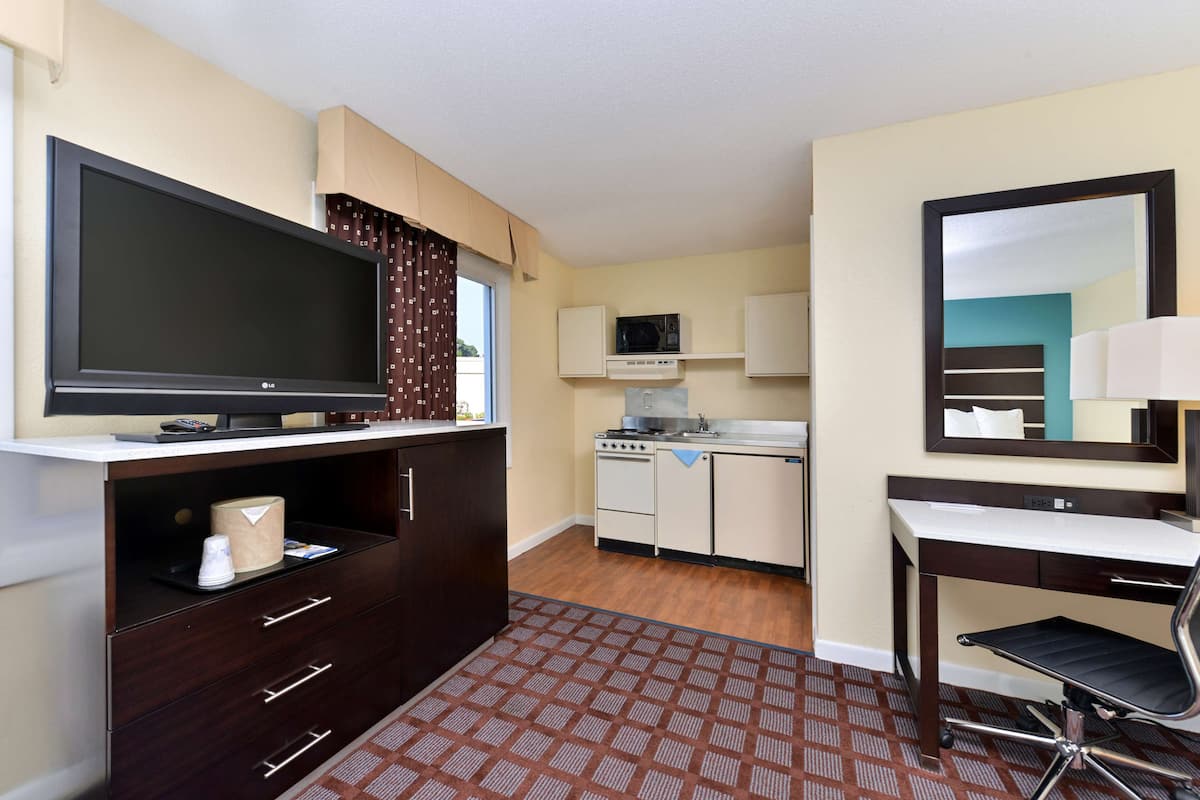 A compact hotel room with a flat-screen TV, a kitchenette, a desk with a chair, and a large mirror.