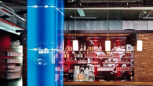 A bar area with shelves of bottles and glasses, a blue pillar, and wooden bar counter. - Cube Savognin (Surses)