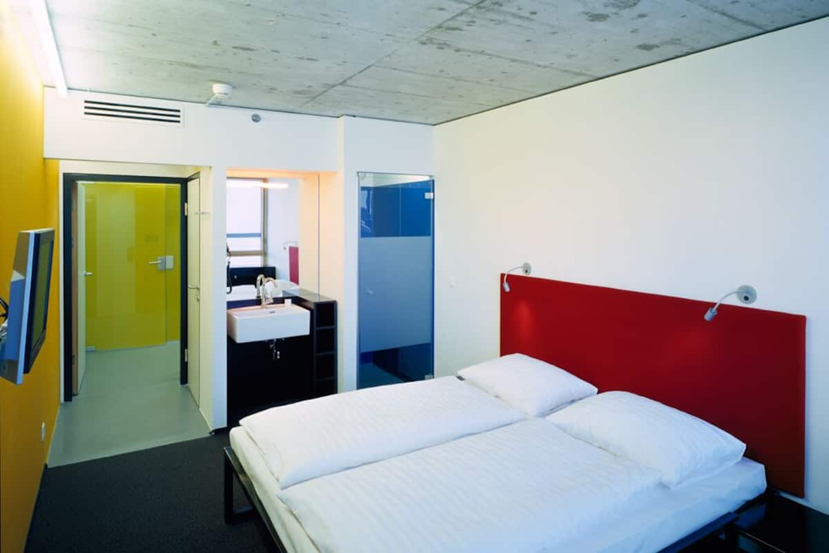 A hotel room with a double bed, a red accent wall, a sink, and a mirror.