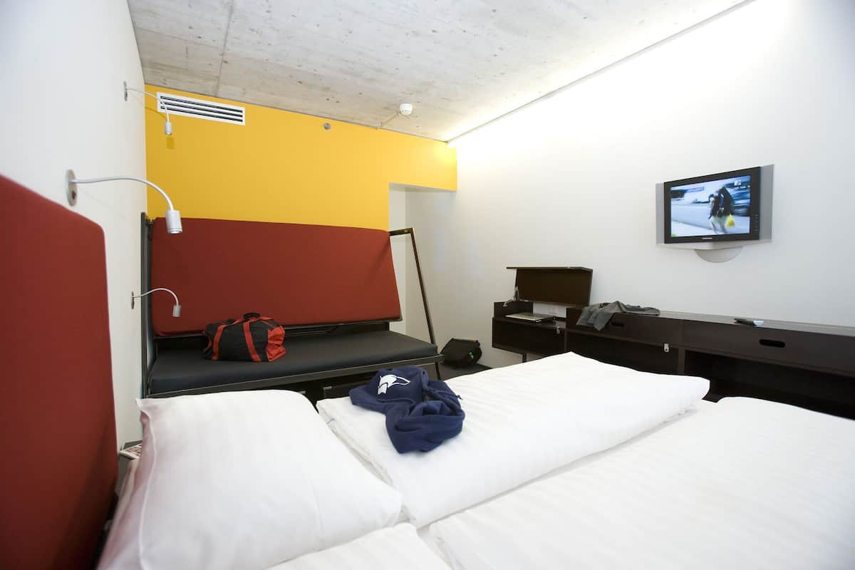 A hotel room with a bed, a desk with a computer, a TV, and a red and yellow wall.