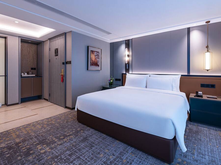 A hotel room with a large bed, a desk, a chair, a nightstand, a wall-mounted lamp, and a door with a handle.