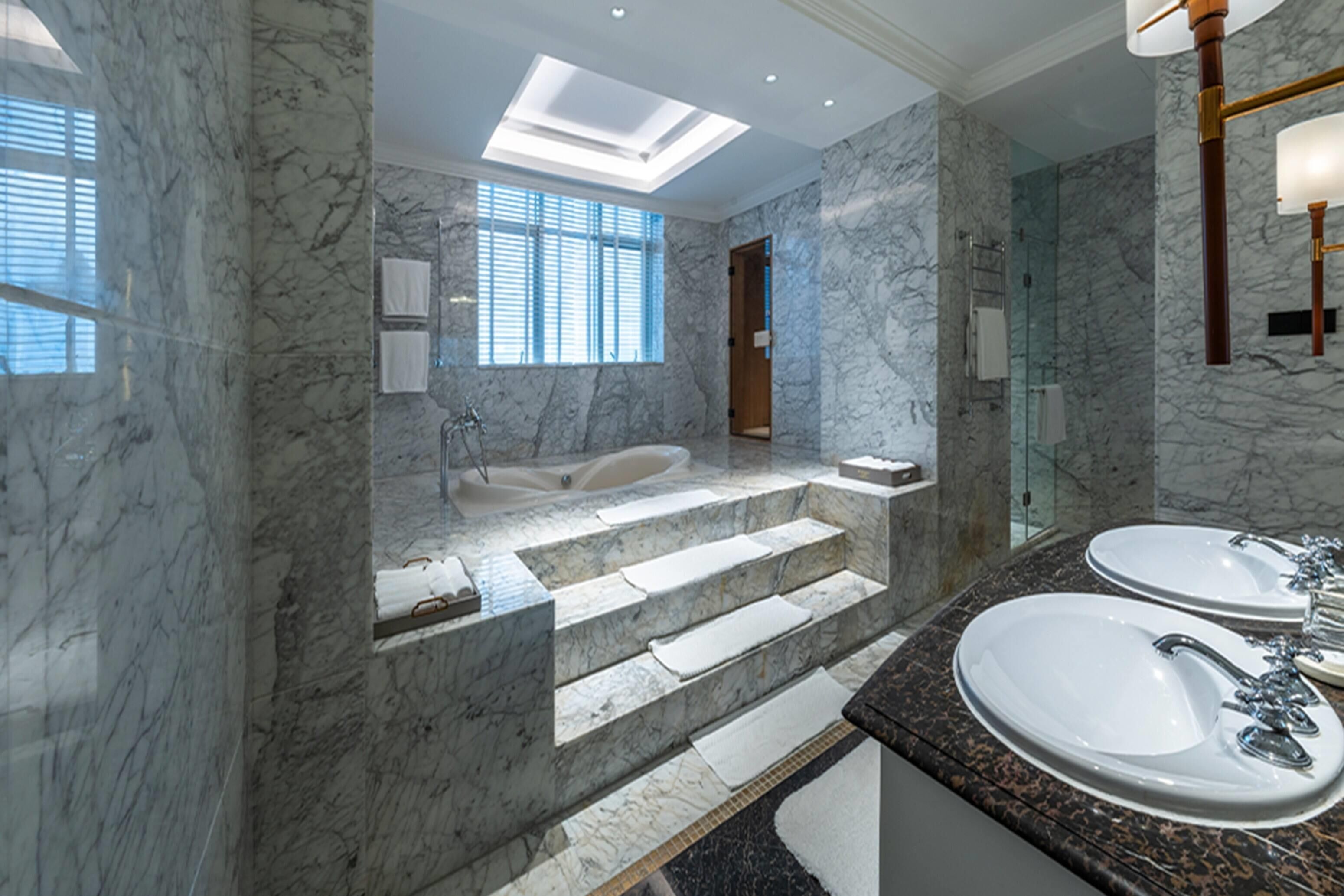 A modern bathroom with marble walls, a skylight, a bathtub, and two sinks.