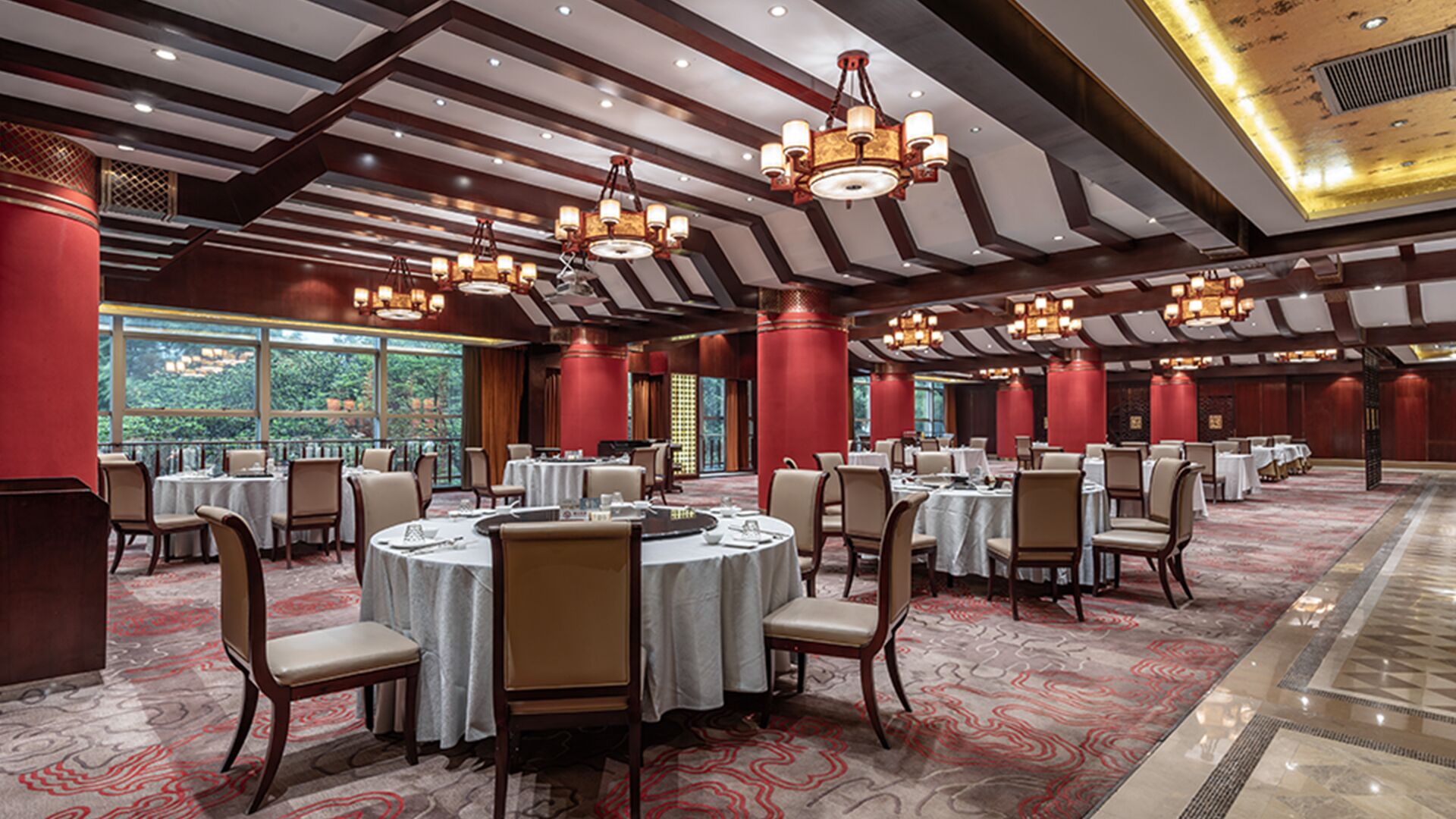 A spacious banquet hall with round tables, red pillars, and chandeliers.