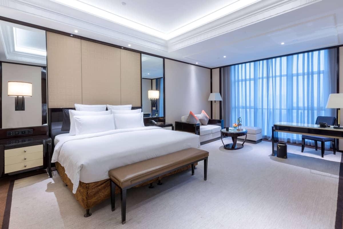 A modern hotel room with a large bed, a desk, a chair, a bench, and a large window with curtains.