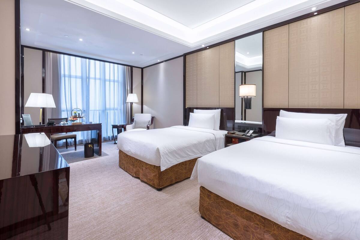 A hotel room with two beds, a desk, a chair, and a large mirror.