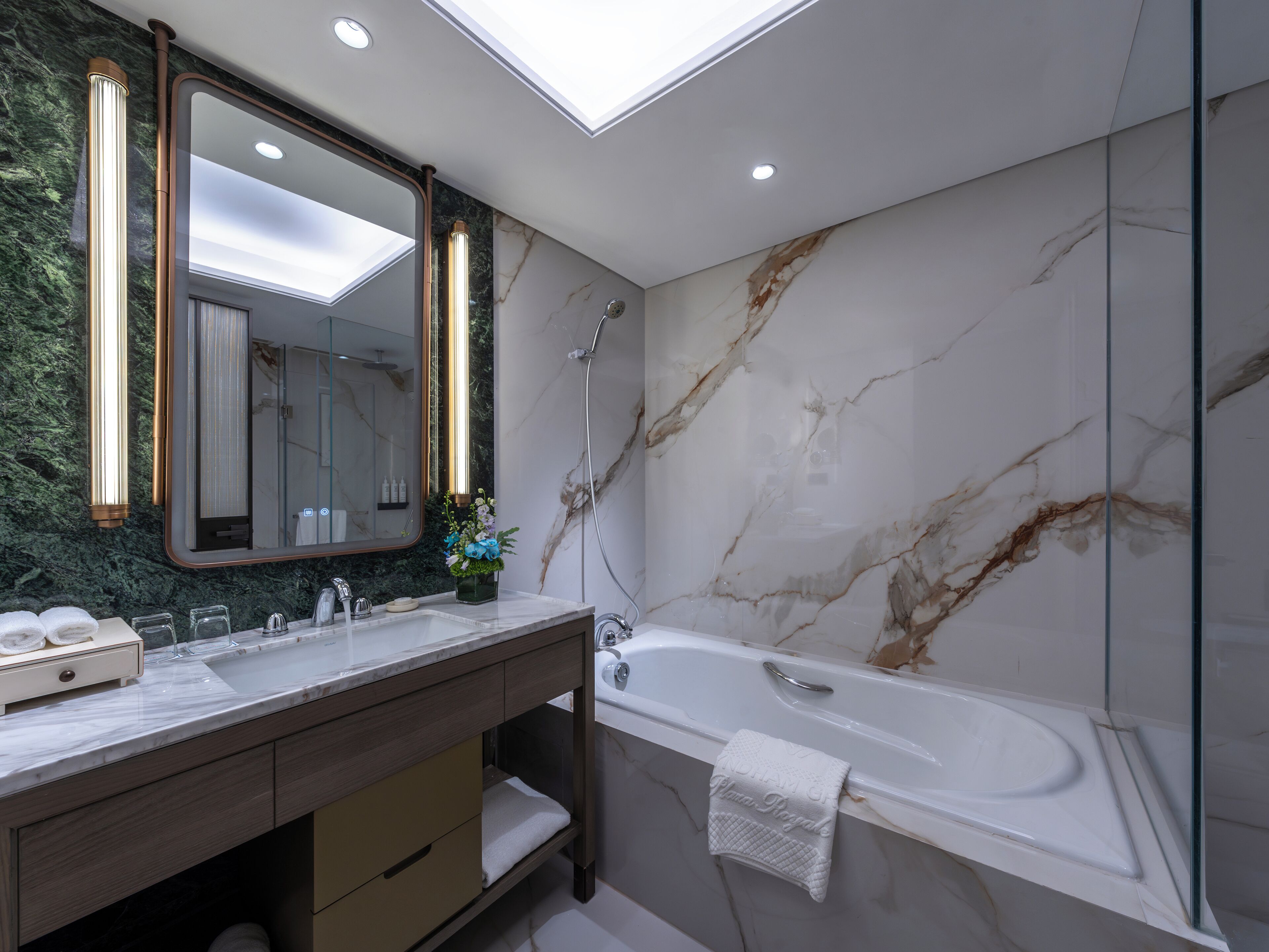 A modern bathroom with a marble wall, a large mirror, a bathtub, and a vanity with a towel rack.