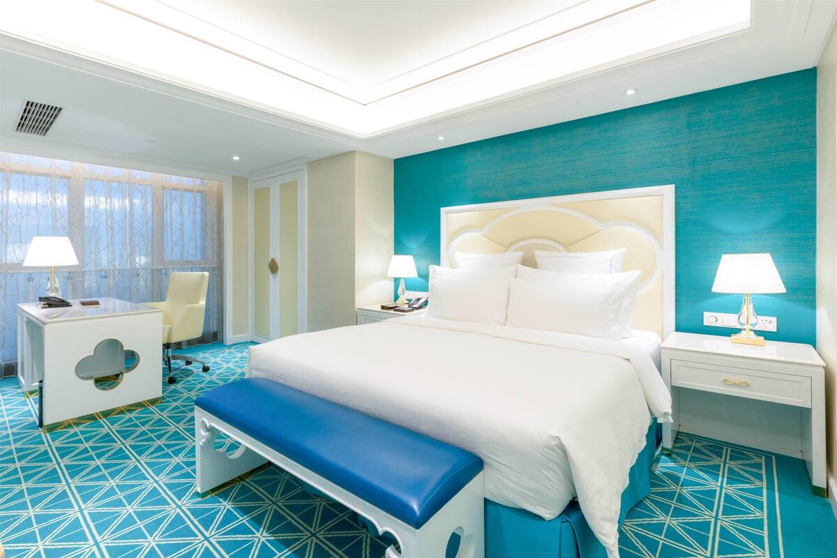 A modern hotel room with a large bed, a bench, a desk, and a chair.