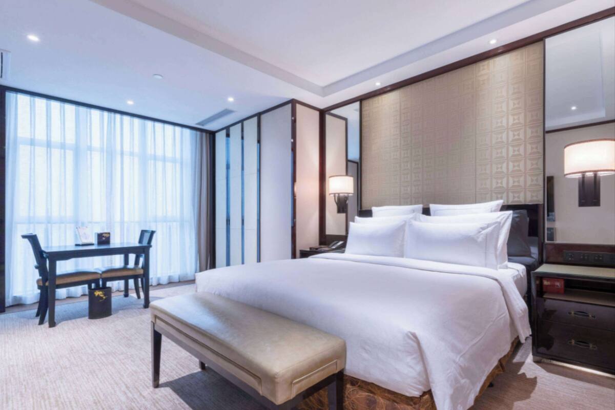 A hotel room with a large bed, a desk with a chair, a bench, and a large window with curtains.