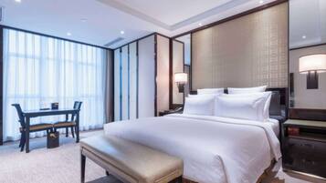 Habitación de hotel con una cama grande, un escritorio con silla, un banco y un ventanal con cortinas.