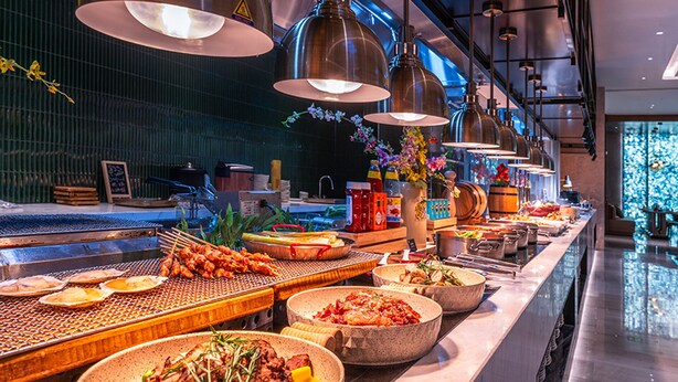 Ein reichhaltiges Buffet mit verschiedenen Speisen, darunter Meeresfrüchte und Salate, in modernem Ambiente mit hängenden Lampen und einer gefliesten Spritzschutzwand.