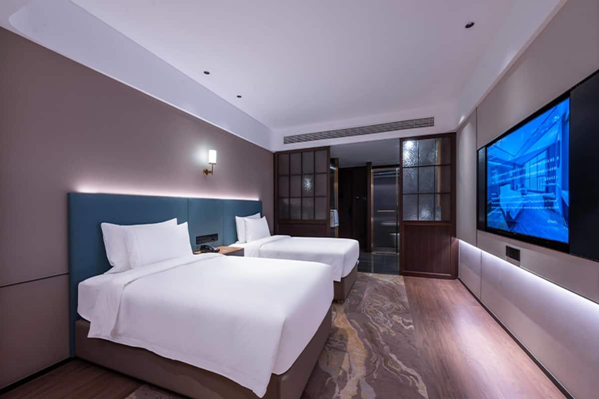 A hotel room with two beds, a large flat-screen TV, and a view of the city through a glass door.
