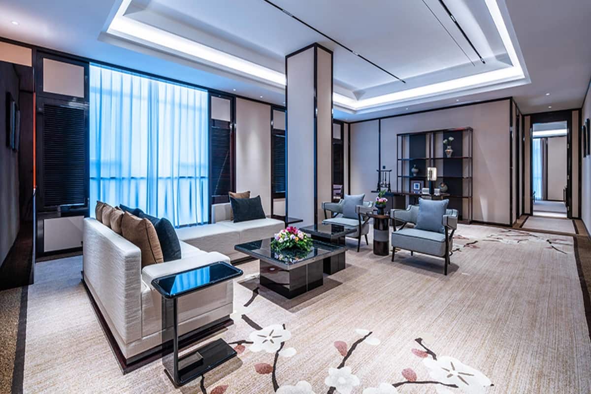 A modern living room with a sofa, coffee table, and armchairs. There is a large window with blinds, a bookshelf, and a decorative vase.
