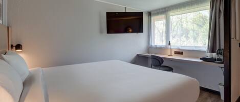 Habitación de hotel con una cama grande, un escritorio con silla, un televisor montado y una ventana con cortinas.
