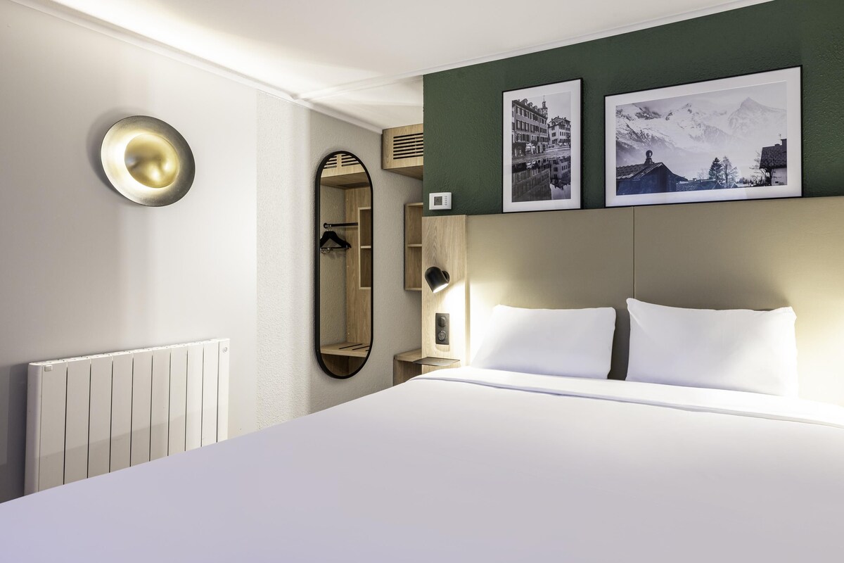 A hotel room with a large bed, a mirror, two framed pictures, a radiator, and a wall-mounted light fixture.