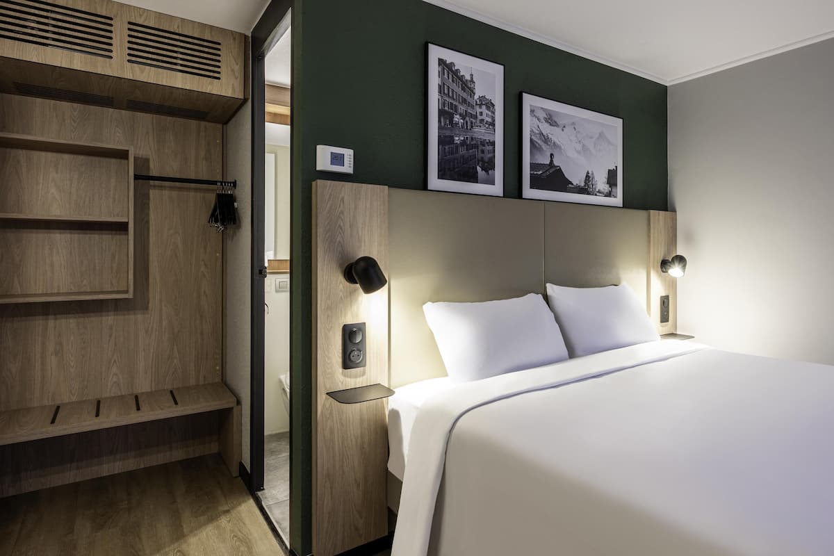 Une chambre d’hôtel moderne avec une armoire en bois, un lit recouvert de draps blancs, deux tableaux encadrés au mur et une table de chevet avec une lampe.