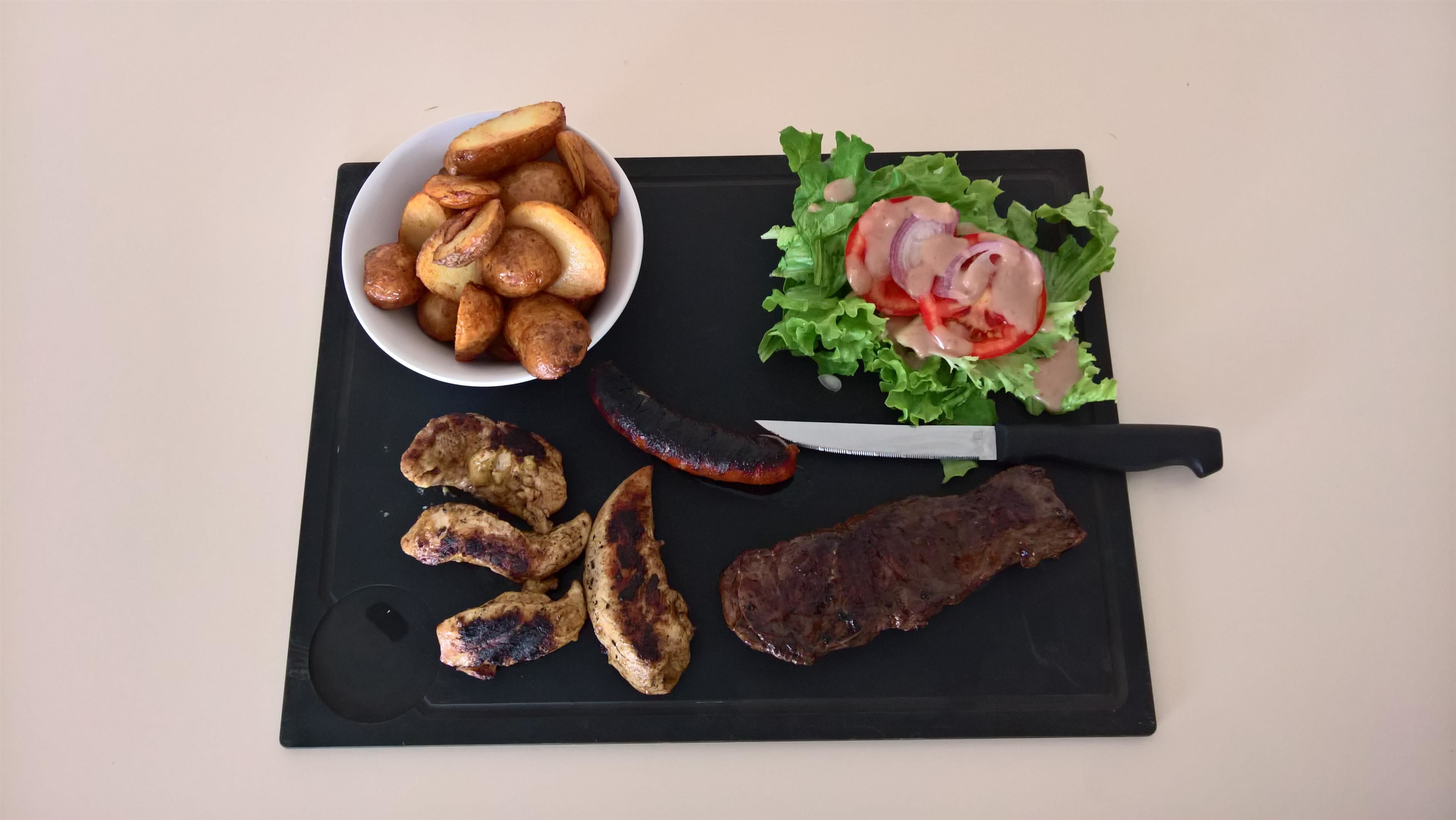 Ein Schneidebrett mit gerösteten Kartoffeln, gegrilltem Hähnchen, Bratwurst und einem Steak.