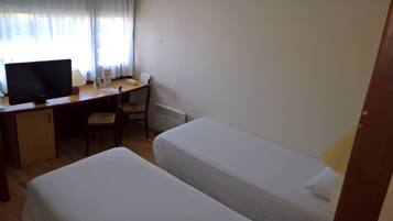 A room with a desk, a computer, two beds, a chair, and a window with curtains.
